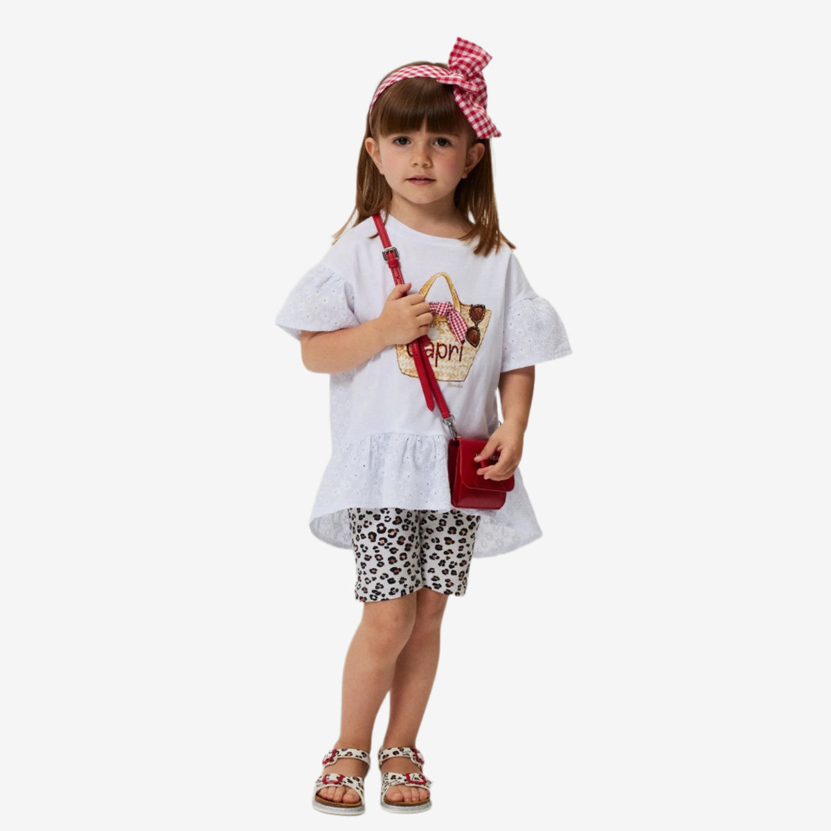 A young girl wearing a white t-shirt with a character print and leopard tulle shorts, accessorized with a red bag and a checkered headband.