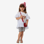 A young girl wearing a white t-shirt with a character print and leopard tulle shorts, accessorized with a red bag and a checkered headband.