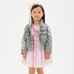 A girl wearing a denim jacket with embroidered flowers, a pink dress, and pink sneakers.