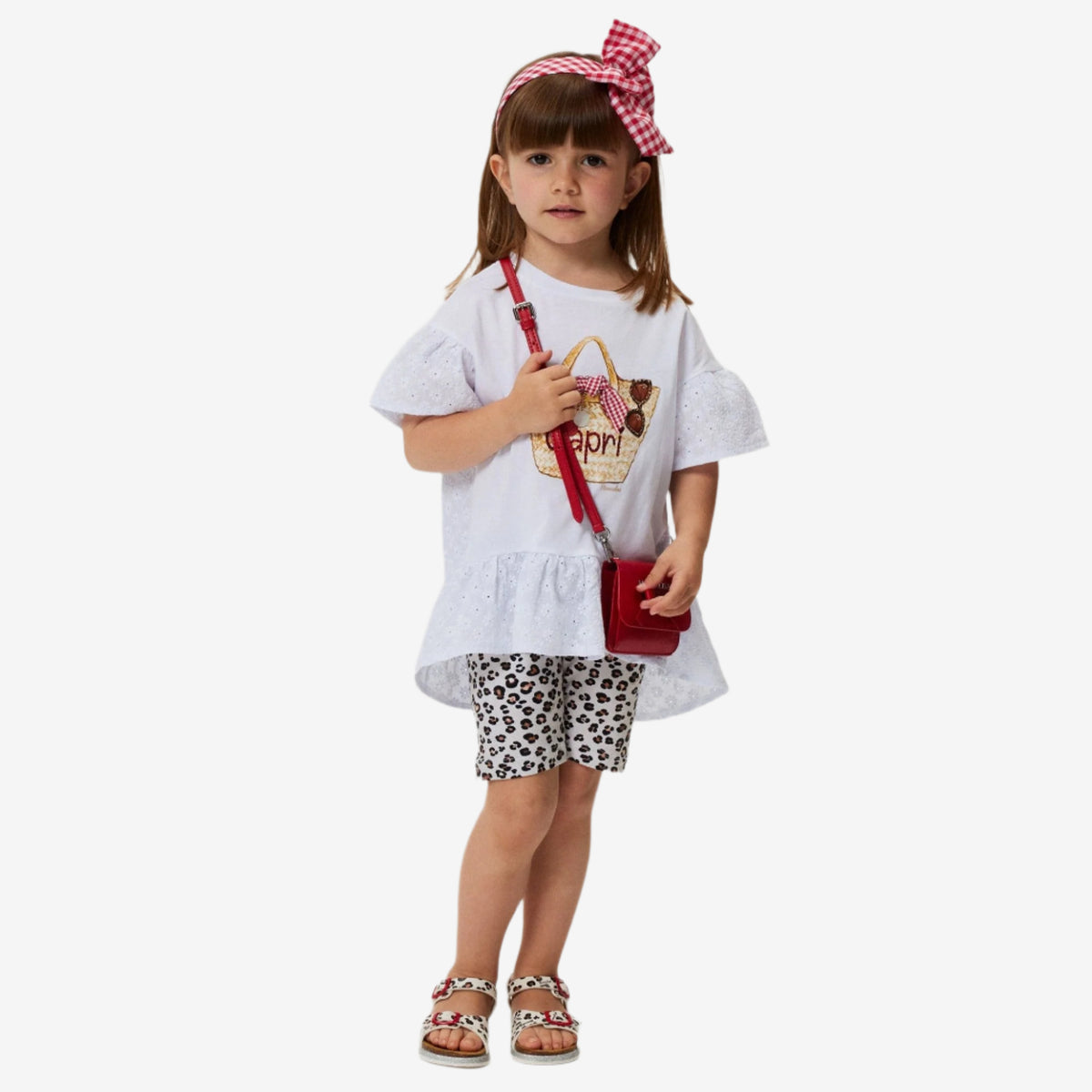 A girl wearing a white maxi t-shirt with a flounce and lace back, paired with animal print shorts and a red bag.