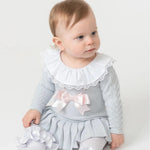 A baby girl wearing a sky blue knitted dress with a lace-trimmed collar and a bow detail on the chest. The dress features a pleated hem.