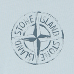 A circular Stone Island compass logo printed in a distressed dark blue-gray tone on a very light icy blue fabric background.