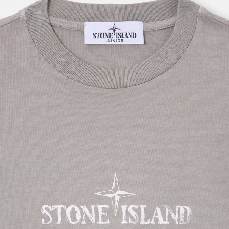 Close-up of a Stone Island Junior t-shirt in a light gray heather color, featuring a white printed compass rose and 'STONE ISLAND' text on the chest, with the brand label visible inside the collar.