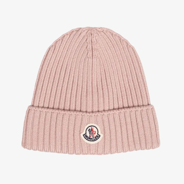 A light pink ribbed knit beanie hat featuring a folded cuff with a circular embroidered Moncler logo patch, set against a solid light gray background.