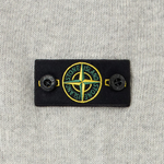 Close-up of a Stone Island woven label patch on a light gray heather knit fabric, featuring the brand's compass logo in yellow, green, and white.