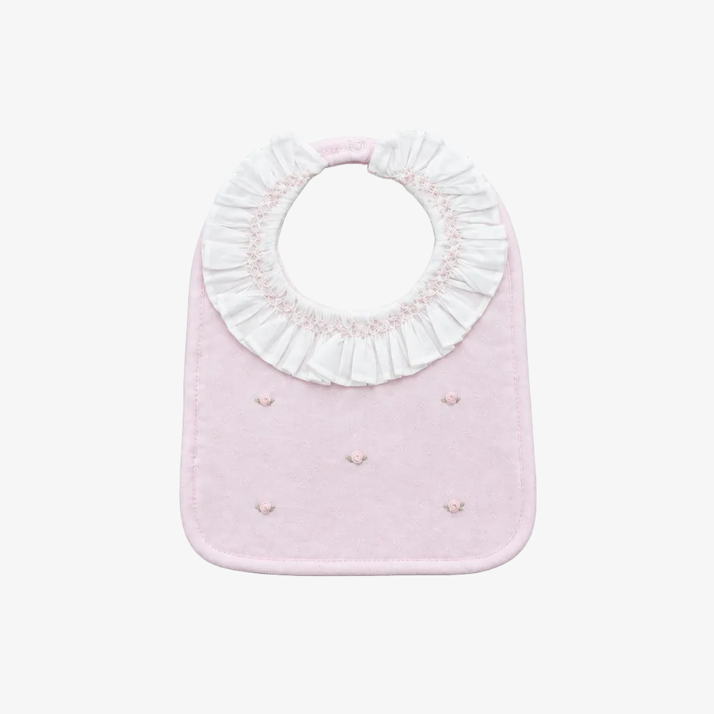 A light pink baby bib with a white ruffled collar and small embroidered details, shown on a neutral gray background.
