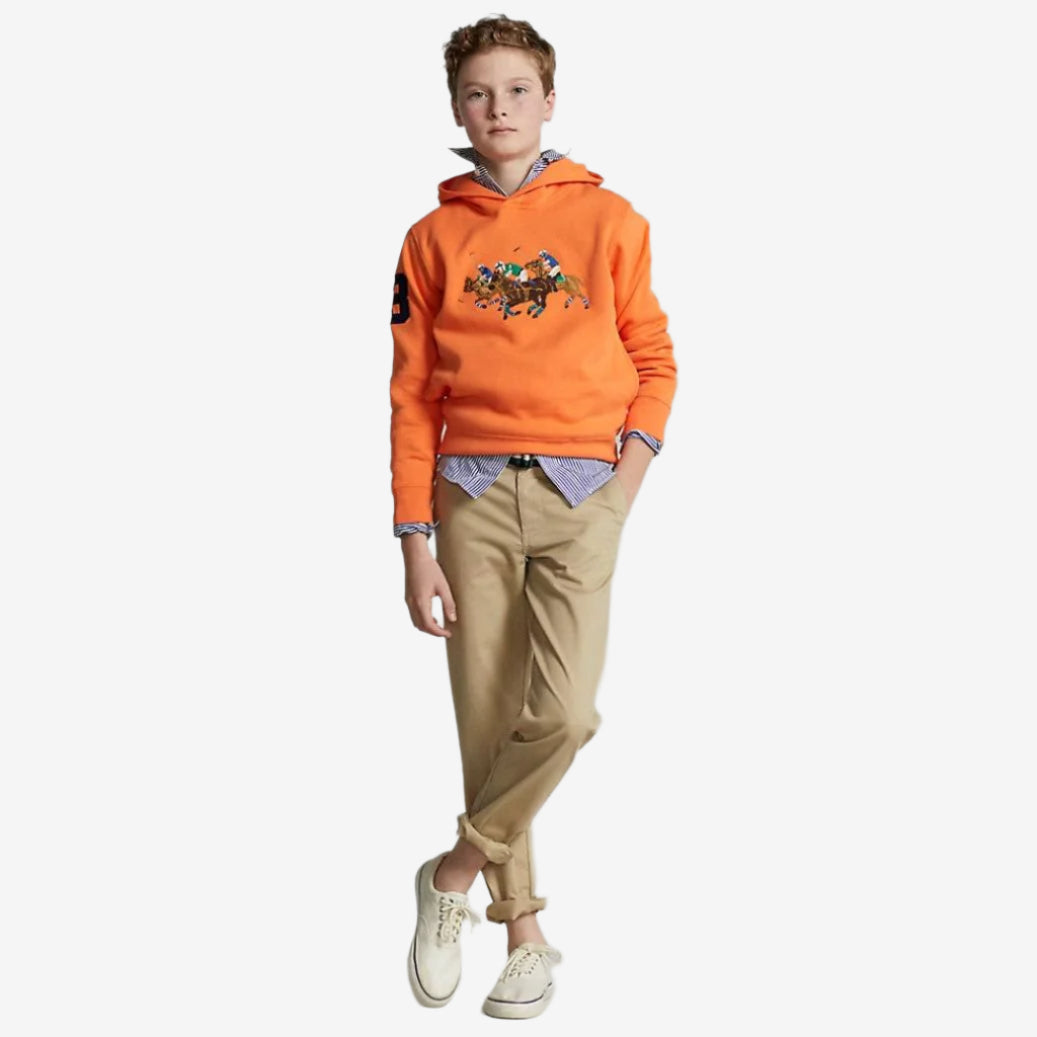 A boy wearing orange chino trousers and a hoodie with a graphic on the front, paired with a blue shirt and white shoes.