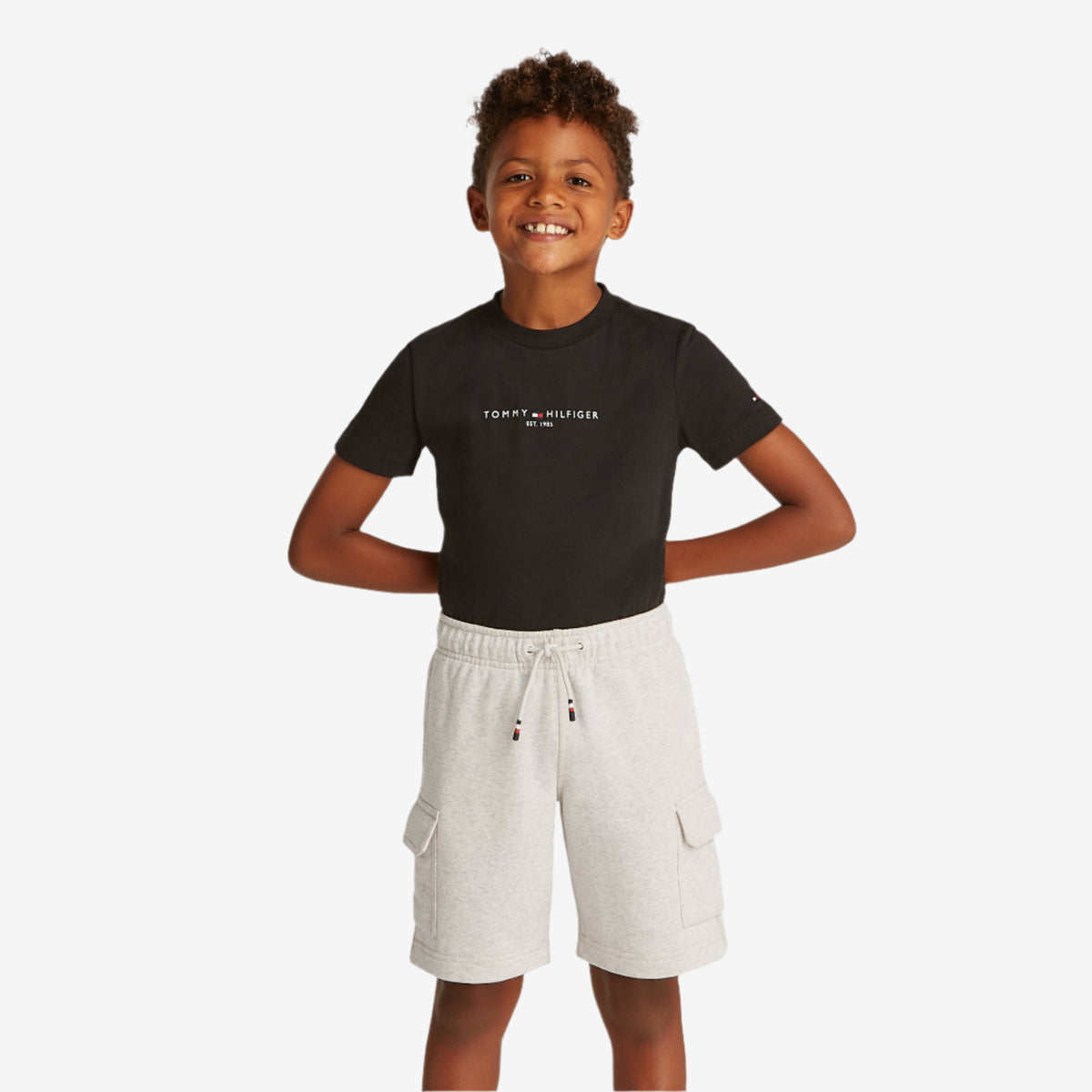 A boy standing and smiling, wearing a black T-shirt and white cargo shorts with a drawstring waist.