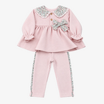 A two-piece toddler girl's outfit set in light pink, featuring a long-sleeve peplum top with a sparkly silver and white patterned Peter Pan collar, a matching bow on the chest, and matching pants with decorative side trim, laid flat on a neutral gray background.