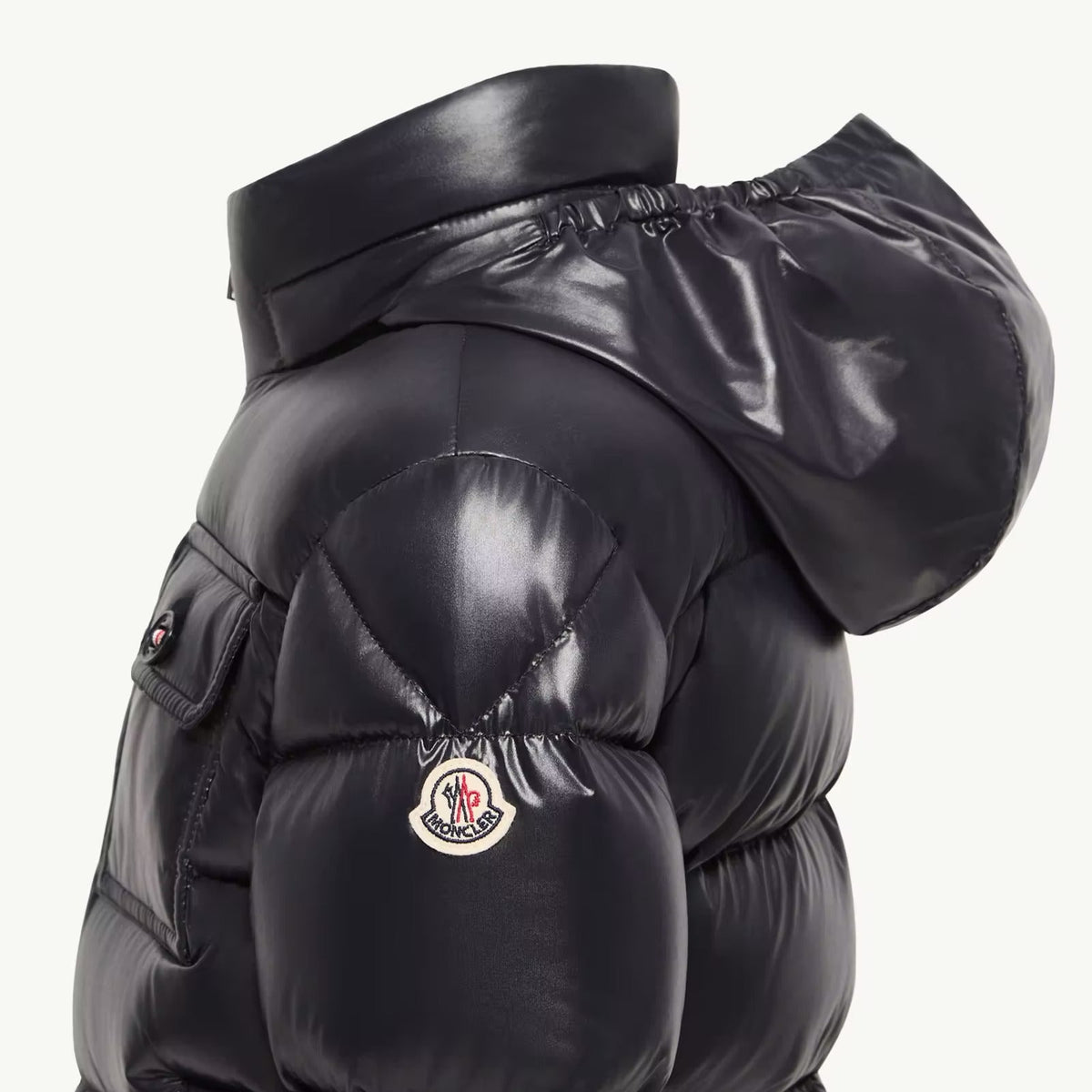 Close-up side view of a glossy black puffer jacket with a high padded collar, quilted sections, and a small circular Moncler logo patch on the upper arm, set against a plain light background.