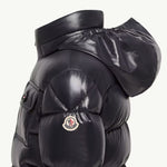 Close-up side view of a glossy black puffer jacket with a high padded collar, quilted sections, and a small circular Moncler logo patch on the upper arm, set against a plain light background.