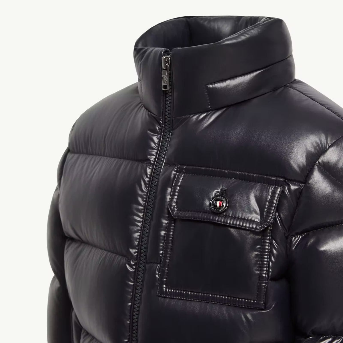 Close-up of a black puffer jacket with a high collar, front zipper, and a chest pocket featuring a small red, white, and blue button detail, shown against a plain light background.