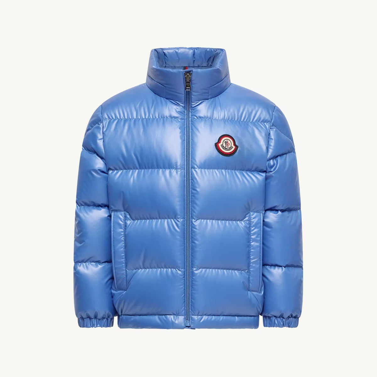 A shiny light blue puffer jacket with a stand-up collar, front zipper, and a circular embroidered Moncler logo patch on the left chest, shown against a plain off-white background.