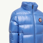 A close-up of a shiny blue puffer jacket with horizontal quilting, a high collar, and the circular red, white, and black Moncler logo patch on the left chest, set against a plain off-white background.