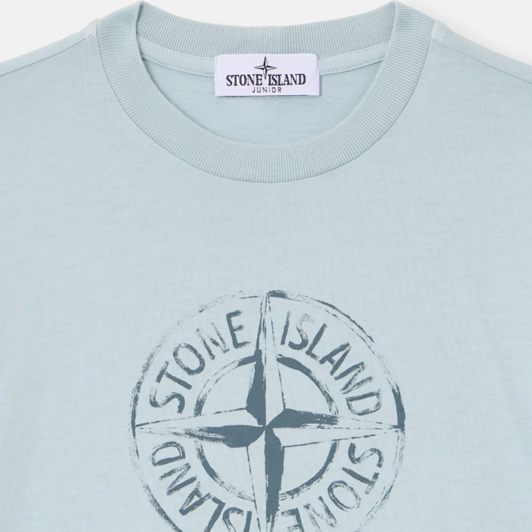 A close-up of a Stone Island t-shirt in a pale icy blue color, featuring the brand's circular compass logo printed in a distressed dark gray on the chest.