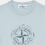 A close-up of a Stone Island t-shirt in a pale icy blue color, featuring the brand's circular compass logo printed in a distressed dark gray on the chest.