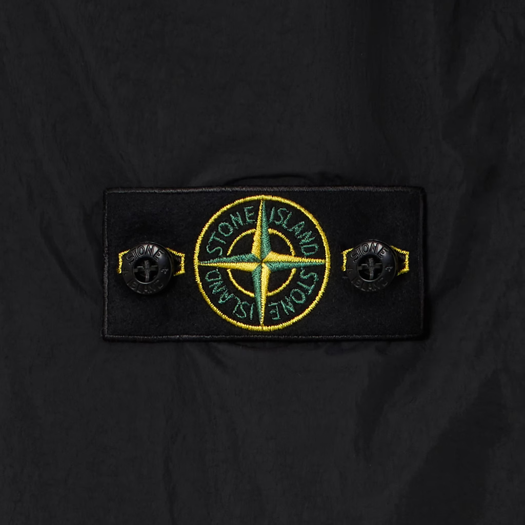 Close-up of a black Stone Island jacket featuring the signature compass badge patch on the chest pocket, flanked by two black buttons.