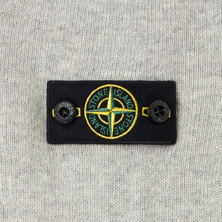 Close-up of a Stone Island woven label patch on a light gray heather knit fabric, featuring the brand's compass logo in yellow, green, and white.