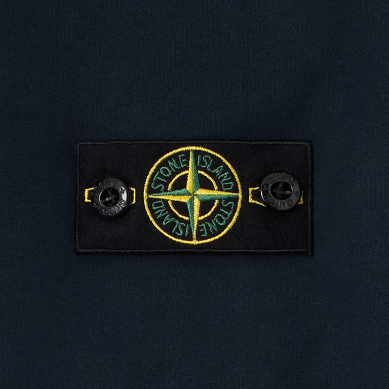 Close-up of a dark navy fabric garment featuring a rectangular black patch with the circular Stone Island compass logo in yellow and green, flanked by two black buttons.