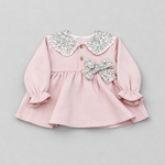 A baby girl's long-sleeve pink dress with a ruffled floral-patterned Peter Pan collar, two gold buttons, and a matching bow on the chest, shown on a neutral gray background.
