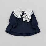 A navy blue long-sleeve baby dress with a large white floral-patterned Peter Pan collar, a matching bow at the center, and ruffled cuffs, laid flat on a light gray background.