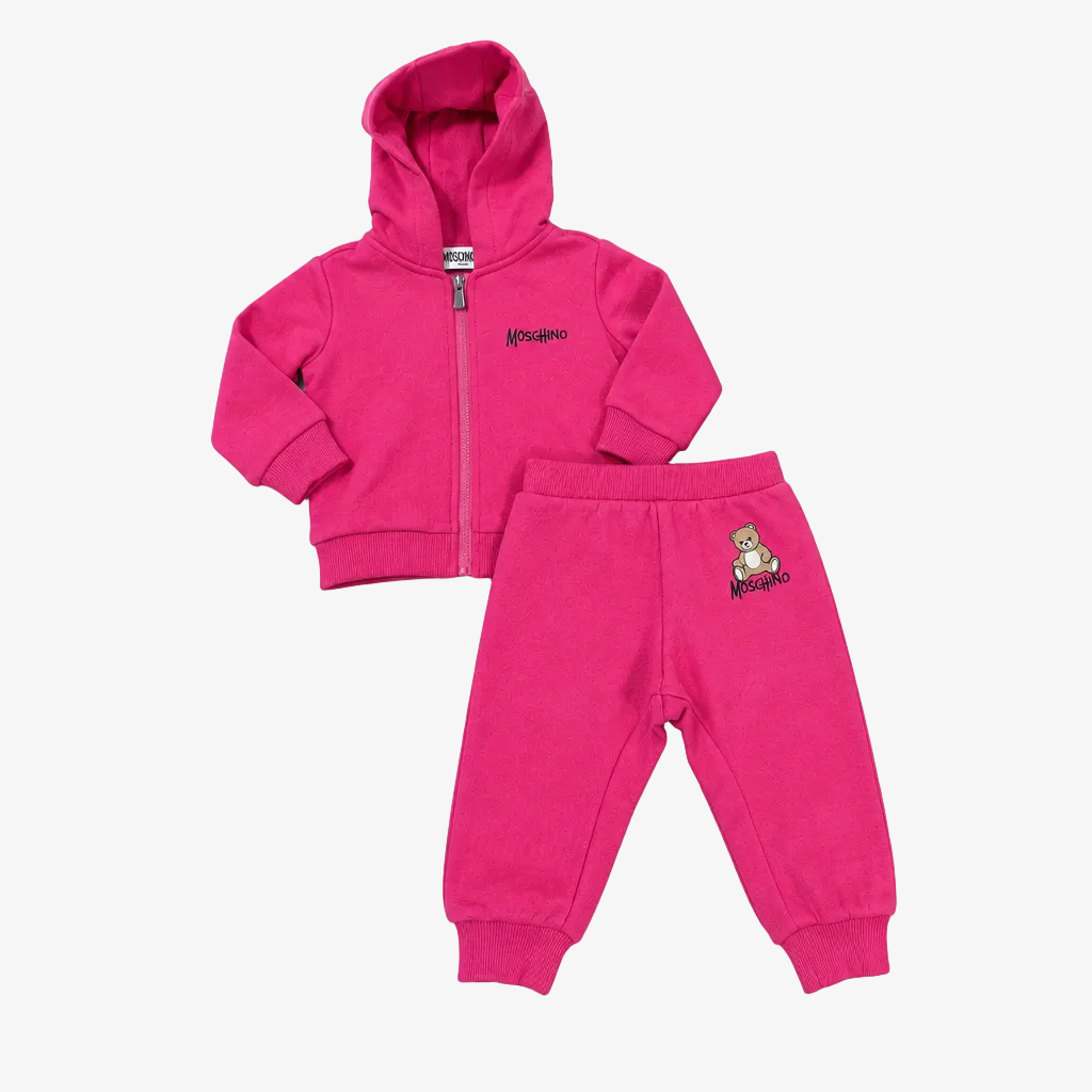 A two-piece kids' outfit set in bright pink, featuring a zip-up hoodie with a small embroidered Moschino logo on the chest and matching jogger pants with a small teddy bear logo on the thigh, laid flat on a light gray background.