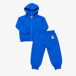 A two-piece kids' tracksuit in a vibrant royal blue, featuring a zip-up hoodie with a small black embroidered logo on the chest and matching jogger pants with a small teddy bear graphic on the thigh. The set is laid flat on a neutral light gray background.