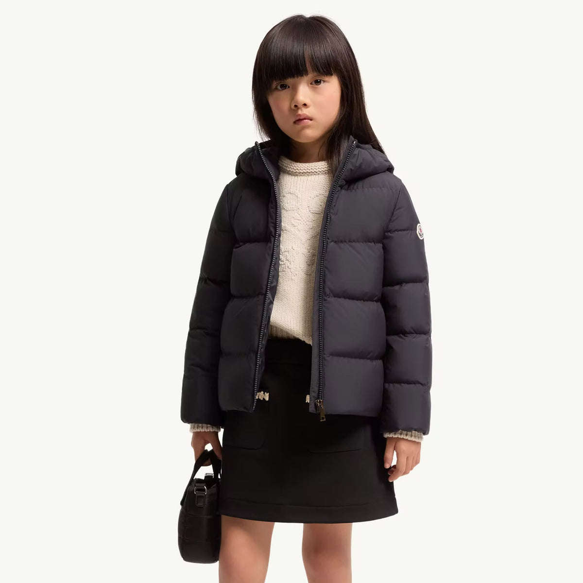 A young girl stands against a plain light background wearing a dark gray puffer jacket over a cream knit sweater, a black skirt, cream socks, and black boots, while holding a small black handbag.