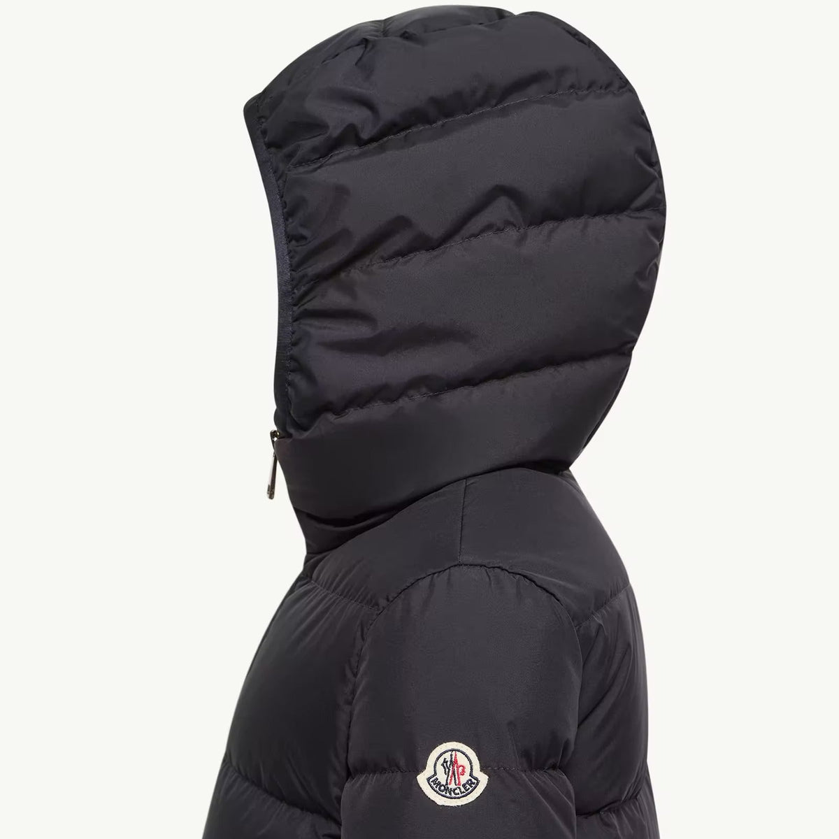 A close-up side view of a black Moncler puffer jacket with the brand's circular logo patch visible on the upper sleeve, set against a plain off-white background.