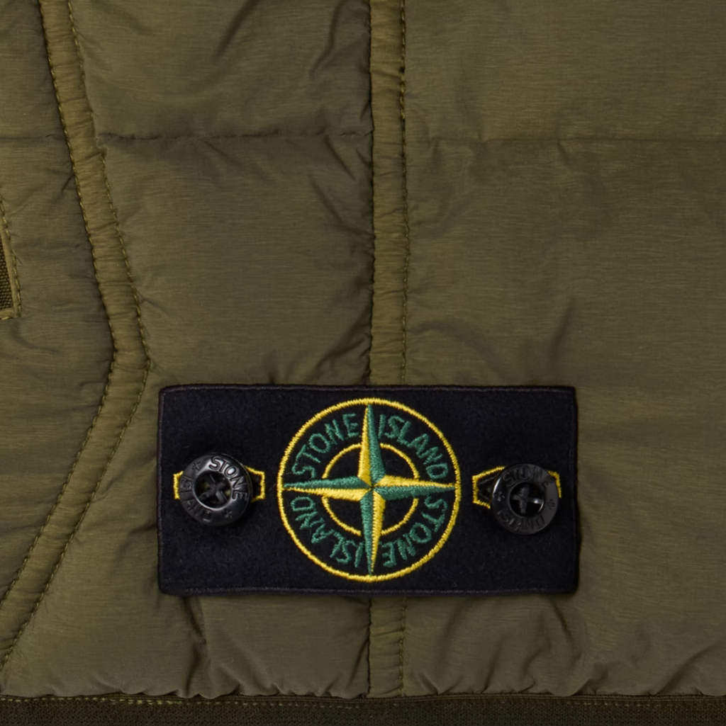 Close-up of an olive green puffer jacket featuring a black rectangular Stone Island logo patch with a yellow and green compass emblem and snap buttons.