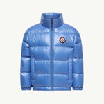 A shiny light blue puffer jacket with a stand-up collar, front zipper, and a circular embroidered Moncler logo patch on the left chest, shown against a plain off-white background.