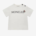 A white short-sleeve baby T-shirt by Moncler, featuring the brand name 'MONCLER' and its signature red bear logo printed on the chest, with two black snap buttons on the shoulder.